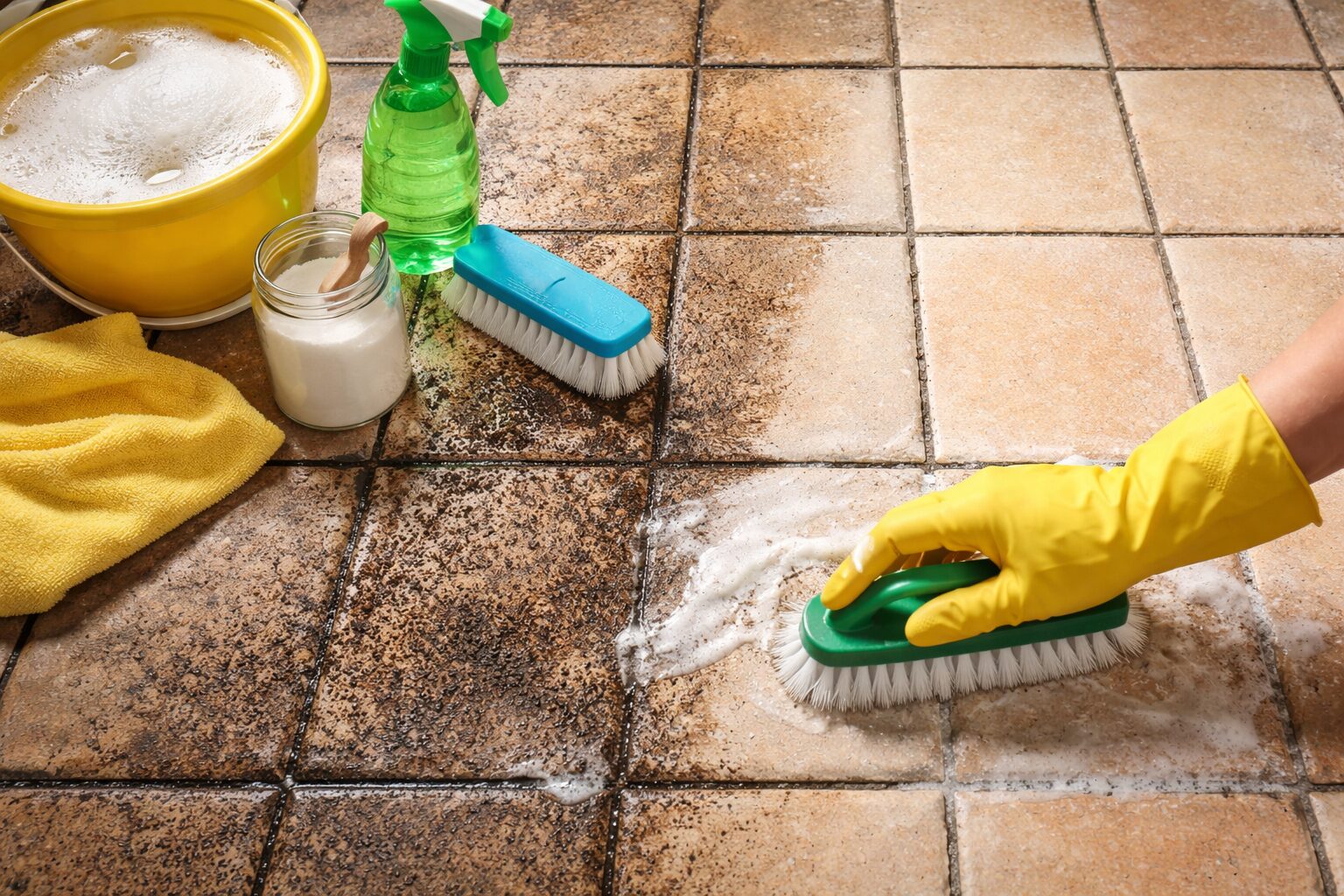 Main avec gant jaune frottant un carrelage poreux très sale, avec des joints foncés, un seau d’eau savonneuse, un spray et une brosse de nettoyage à côté