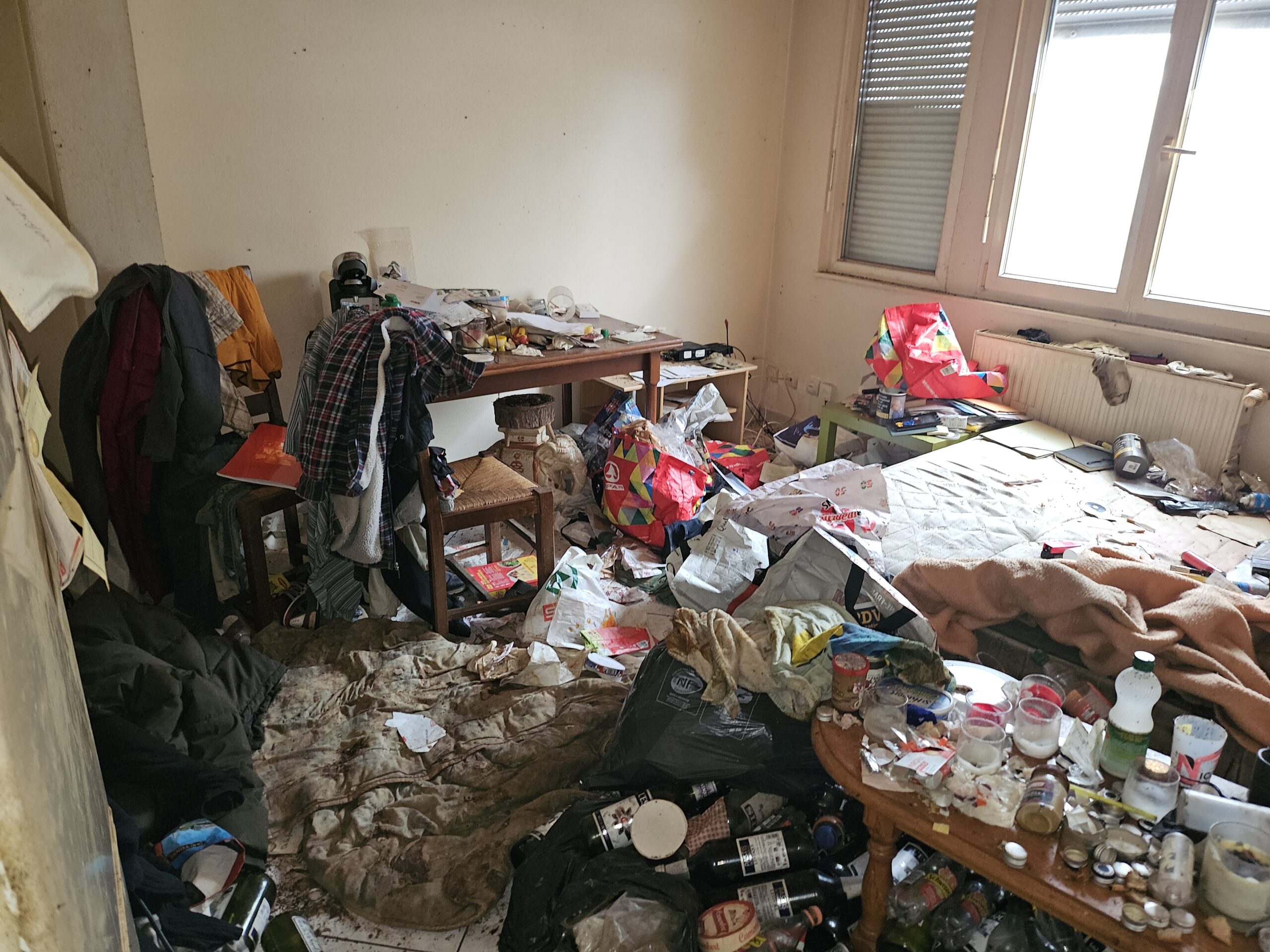 Chambre très sale et encombrée avec déchets, vêtements, bouteilles et objets éparpillés sur le sol et les meubles.