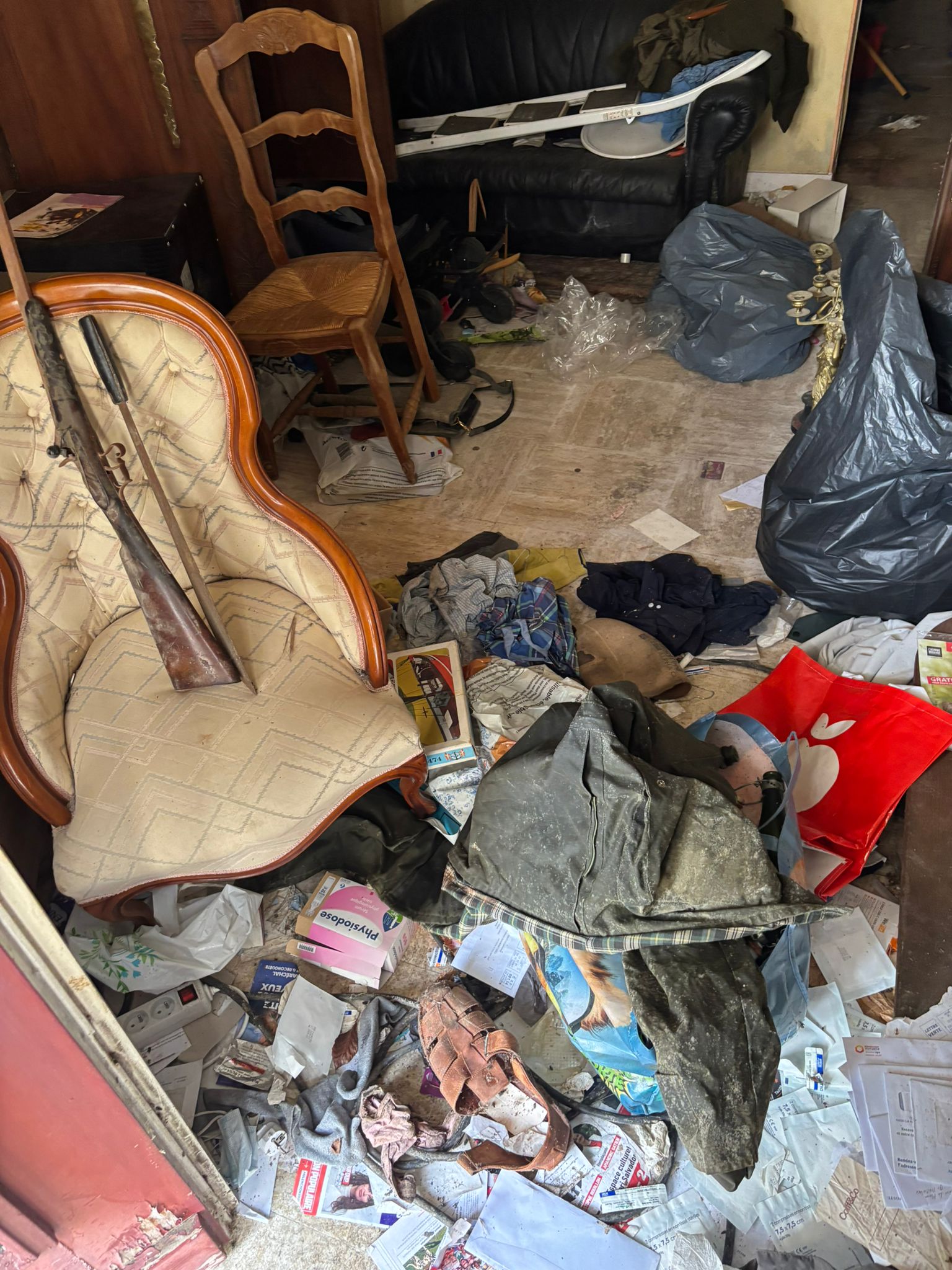 Intérieur d’une pièce en désordre avec meubles anciens, vêtements, papiers et objets divers dispersés au sol.