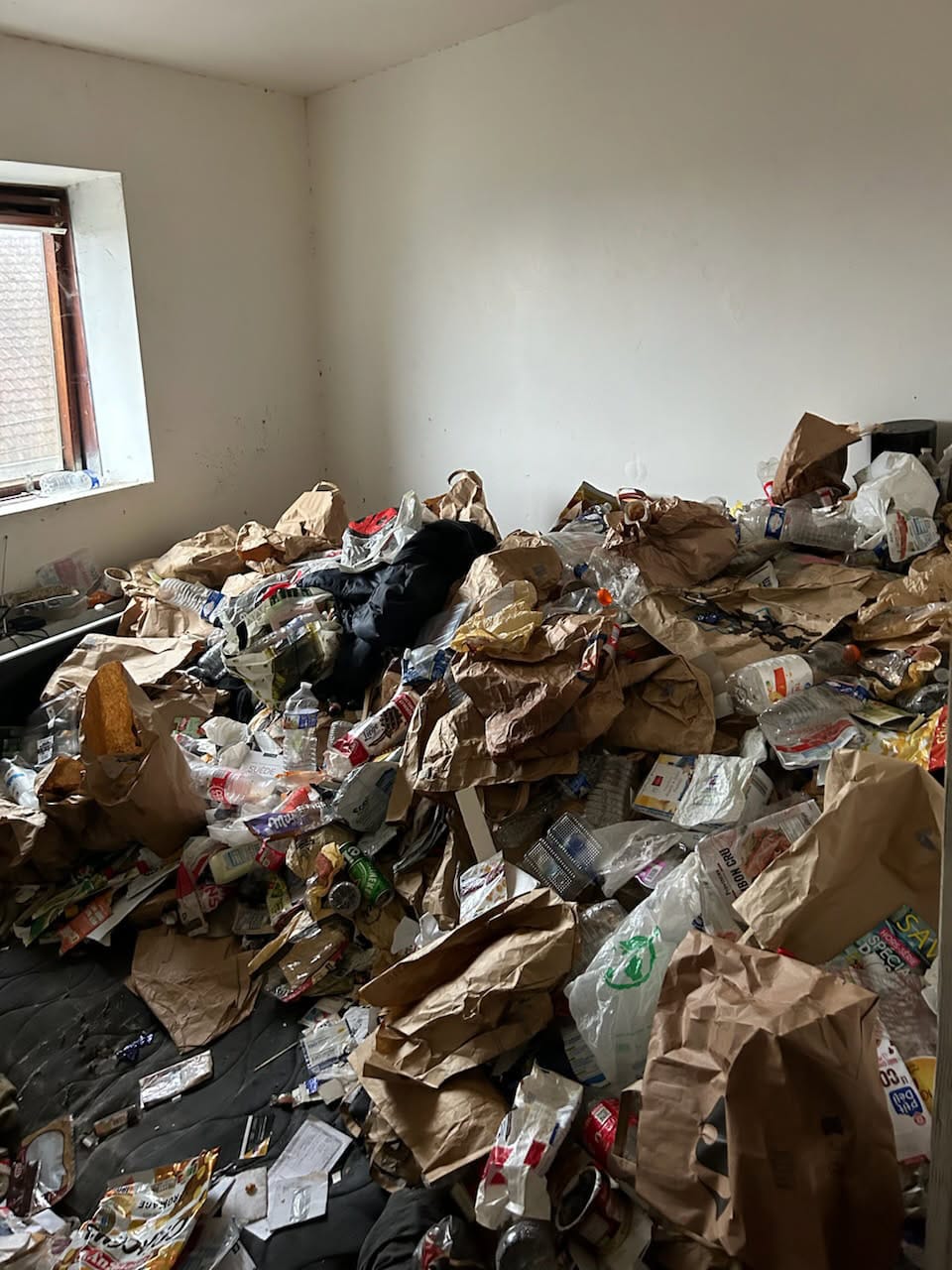 Pièce très encombrée avec accumulation de sacs en papier, bouteilles plastiques, emballages et déchets au sol, dans un contexte de logement insalubre suite à un syndrome de diogène