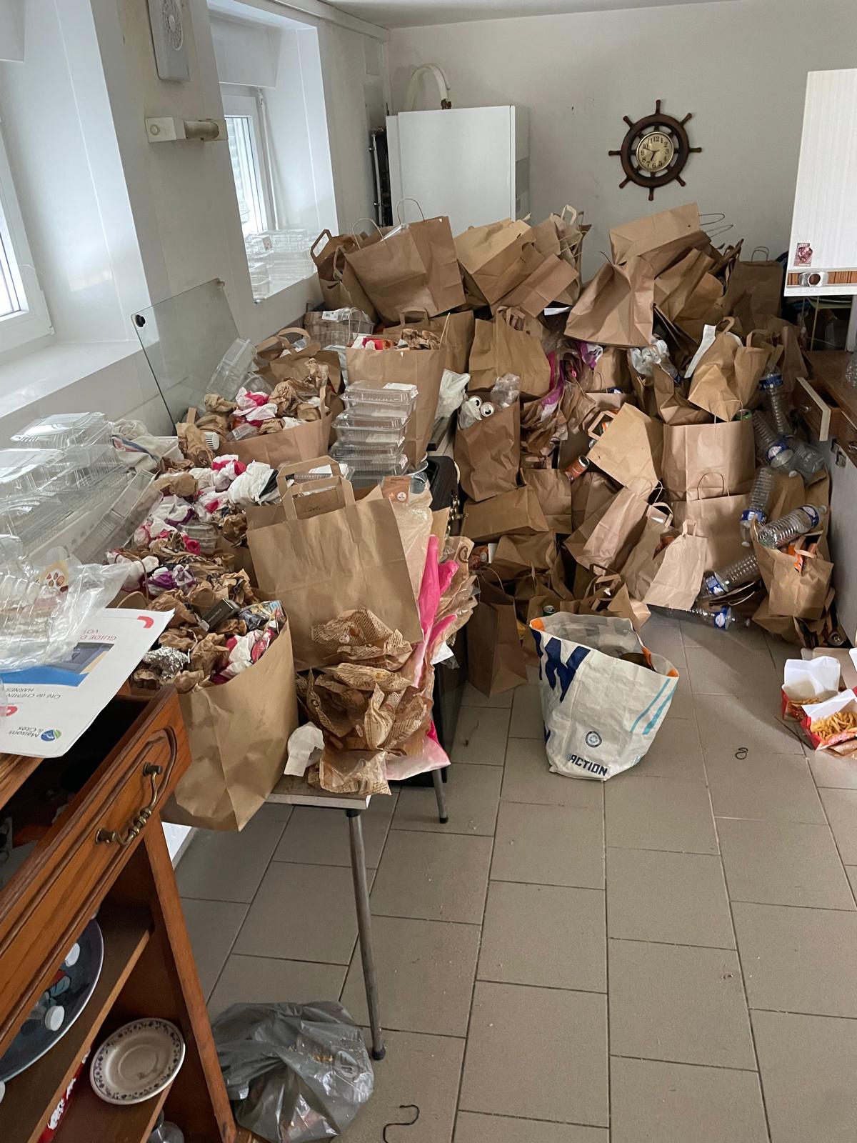 Pièce intérieure très encombrée avec nombreux sacs en papier, emballages alimentaires, bouteilles plastiques et déchets accumulés au sol et sur le mobilier.
