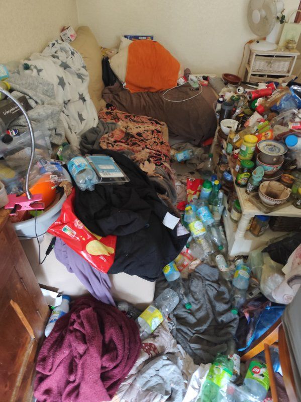 Petite pièce très encombrée avec lit ou canapé recouvert d’objets, bouteilles plastiques au sol, table chargée de contenants et déchets dispersés dans l’espace.