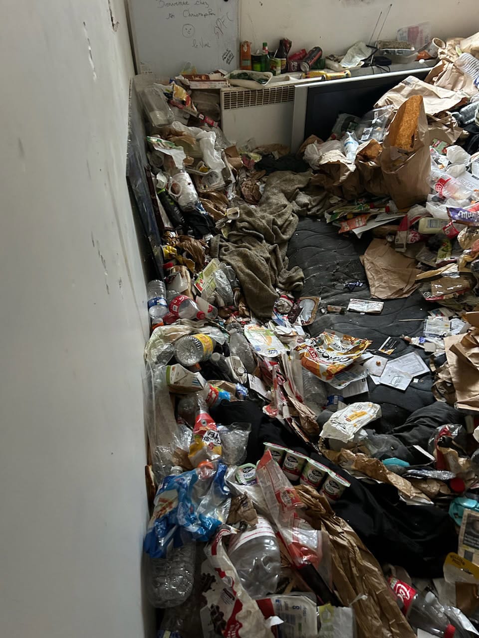 Pièce intérieure encombrée avec matelas au sol recouvert de déchets, bouteilles plastiques, emballages alimentaires et sacs accumulés le long des murs.