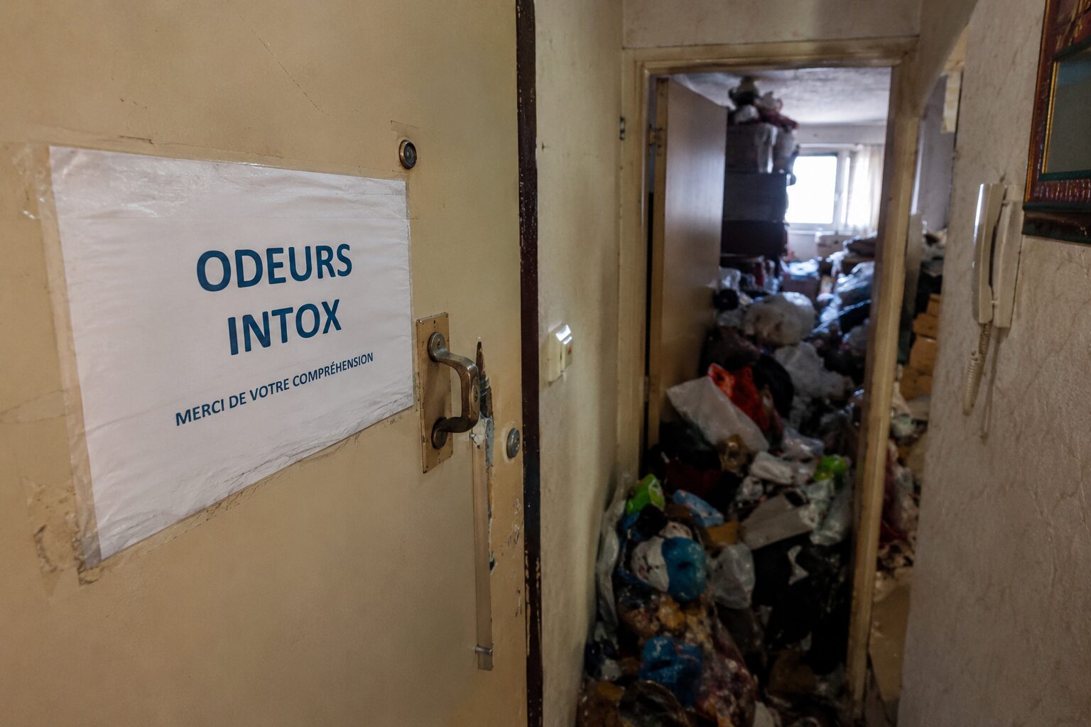 Cage d’escalier d’immeuble avec porte ouverte sur un logement très encombré provoquant des odeurs fortes dans les parties communes