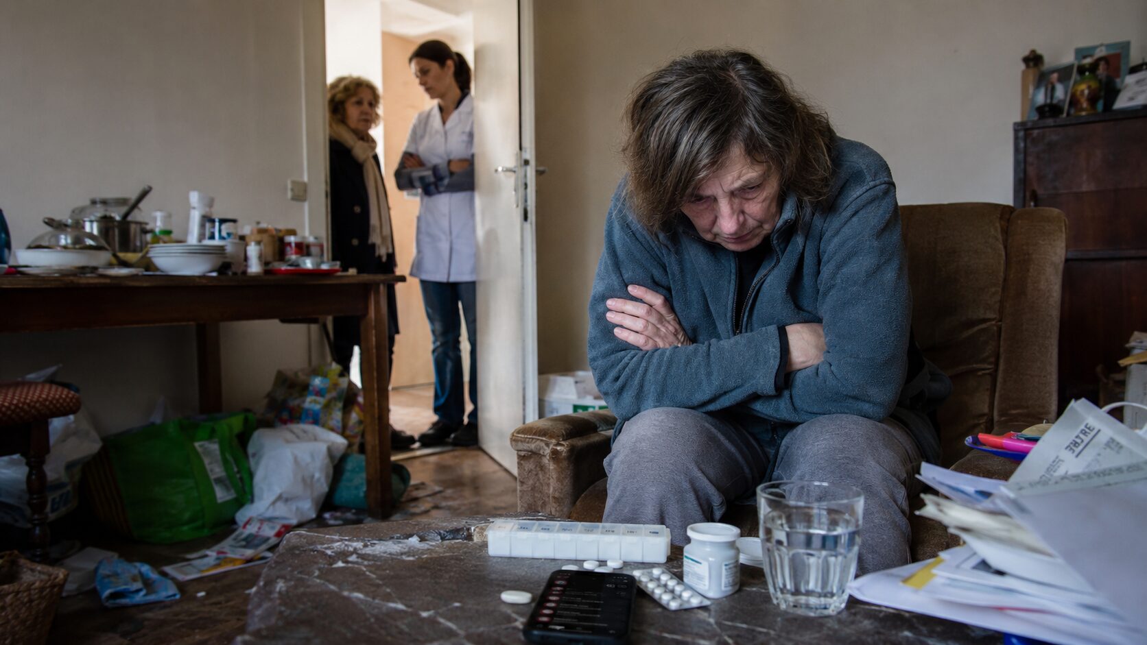 Personne en situation d’incurie assise dans un logement négligé, avec des médicaments sur la table et des proches accompagnés d’une soignante à la porte