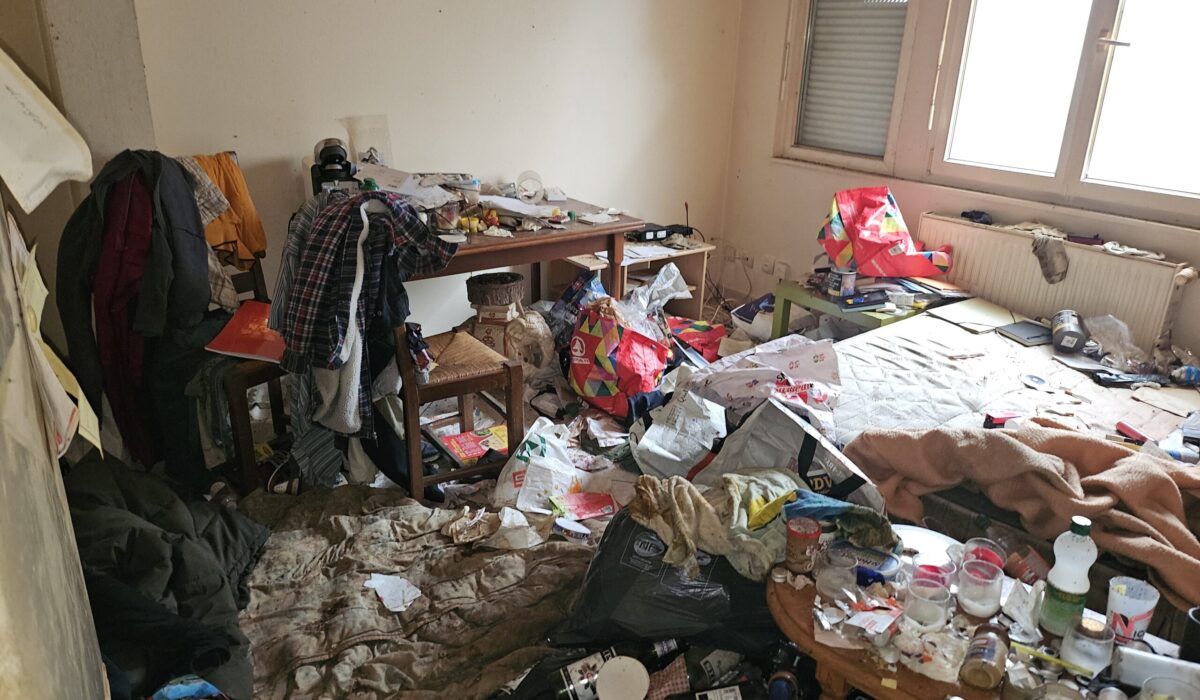 Chambre très sale et encombrée avec déchets, vêtements, bouteilles et objets éparpillés sur le sol et les meubles.