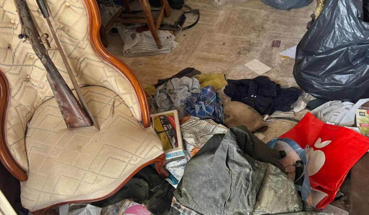 Intérieur d’une pièce en désordre avec meubles anciens, vêtements, papiers et objets divers dispersés au sol.