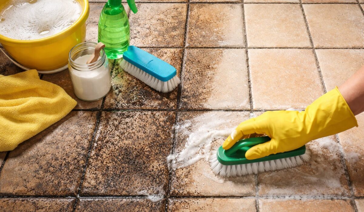 Main avec gant jaune frottant un carrelage poreux très sale, avec des joints foncés, un seau d’eau savonneuse, un spray et une brosse de nettoyage à côté