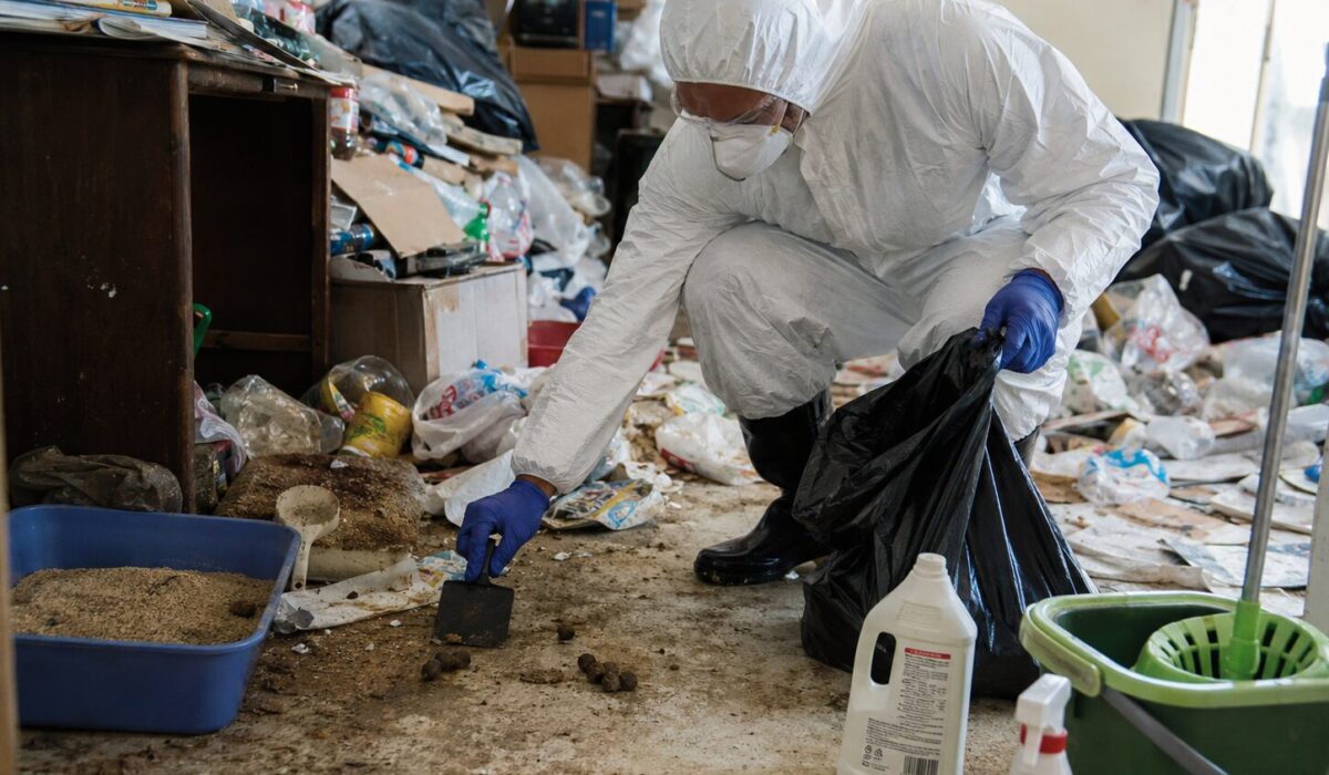 Technicien équipé d’une combinaison de protection nettoyant des excréments d’animaux mélangés à des déchets ménagers dans un logement Diogène insalubre.