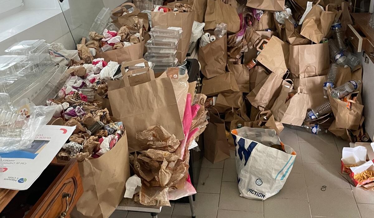 Pièce intérieure très encombrée avec nombreux sacs en papier, emballages alimentaires, bouteilles plastiques et déchets accumulés au sol et sur le mobilier.