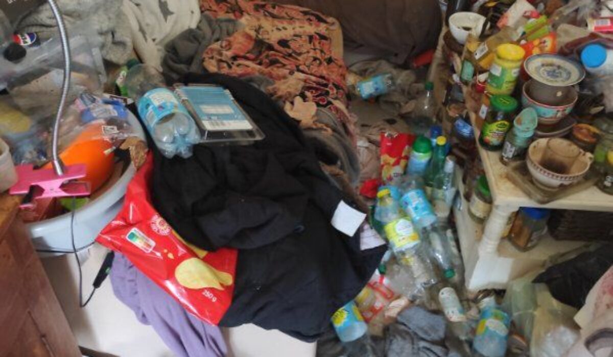 Petite pièce très encombrée avec lit ou canapé recouvert d’objets, bouteilles plastiques au sol, table chargée de contenants et déchets dispersés dans l’espace.