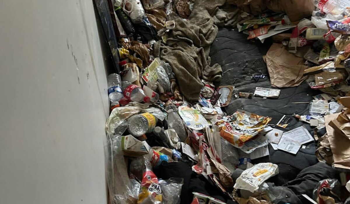 Pièce intérieure encombrée avec matelas au sol recouvert de déchets, bouteilles plastiques, emballages alimentaires et sacs accumulés le long des murs.
