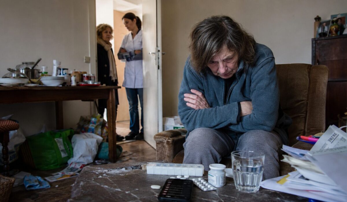 Personne en situation d’incurie assise dans un logement négligé, avec des médicaments sur la table et des proches accompagnés d’une soignante à la porte